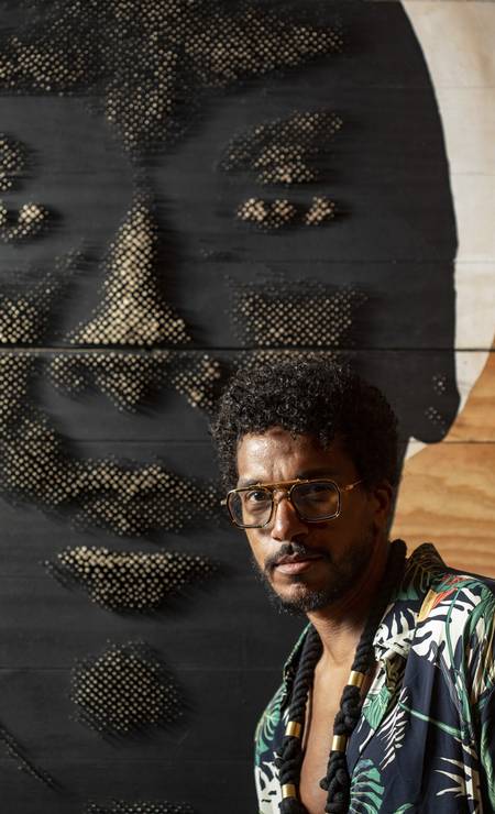 O artista Alexis Peskine - homem negro, usando óculos e camista colorida- em pé ao lado de uma pintura