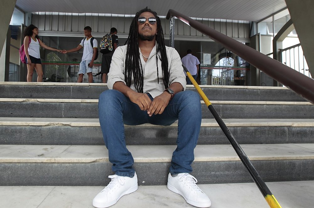 Miguel - homem negro de dreads, usando camiseta e calça jeans - sentado em uma escada