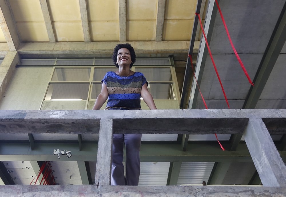 Cássia - mulher negra de cabelo crespo, usando camiseta colorida- em pé sorrindo