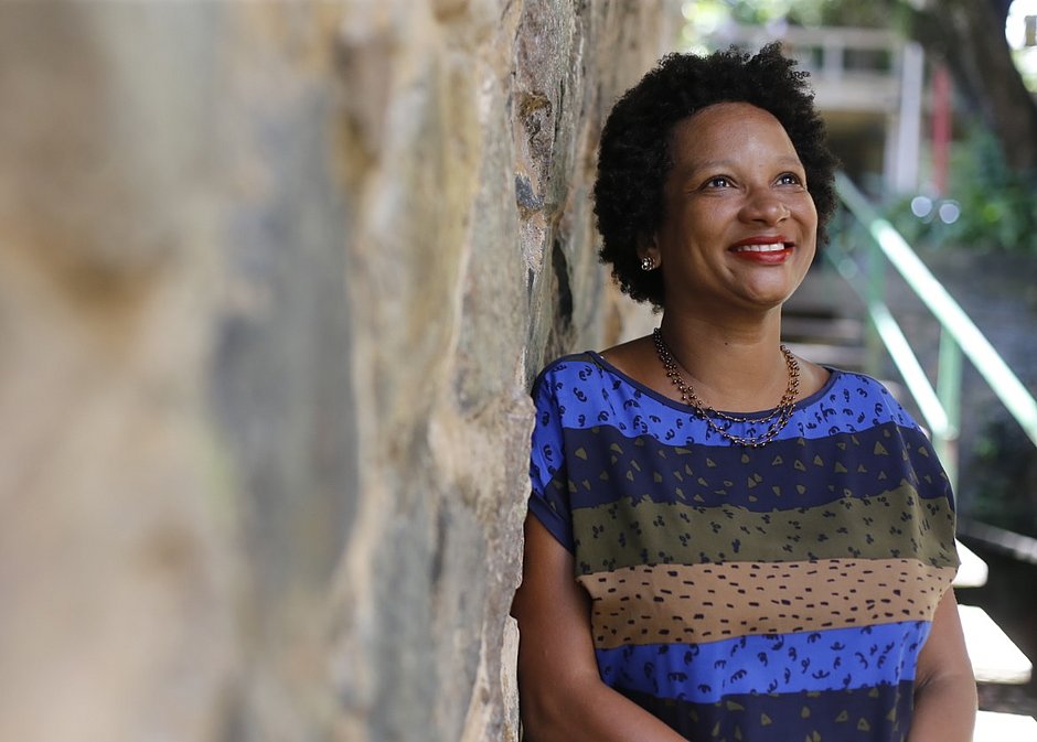 Cássia Maciel - mulher negra de cabelo curto e crespo, usando camiseta colorida- em pé olhando para cima e sorrindo