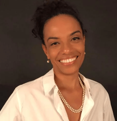 Flávia Oliveira- mulher negra, de cabelo preso, usando camiseta branca - em pé sorrido
